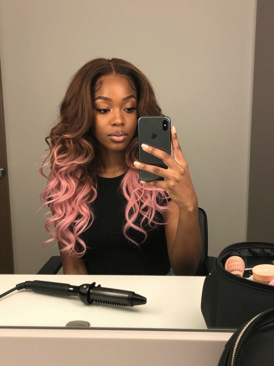 Photo of a black woman wearing soft brown hair with pastel pink tips in loose curls, candid mirror selfie, bathroom vanity mirror setting, soft indoor lighting, with a curling iron on the counter and a makeup bag nearby, latest iPhone photo quality.