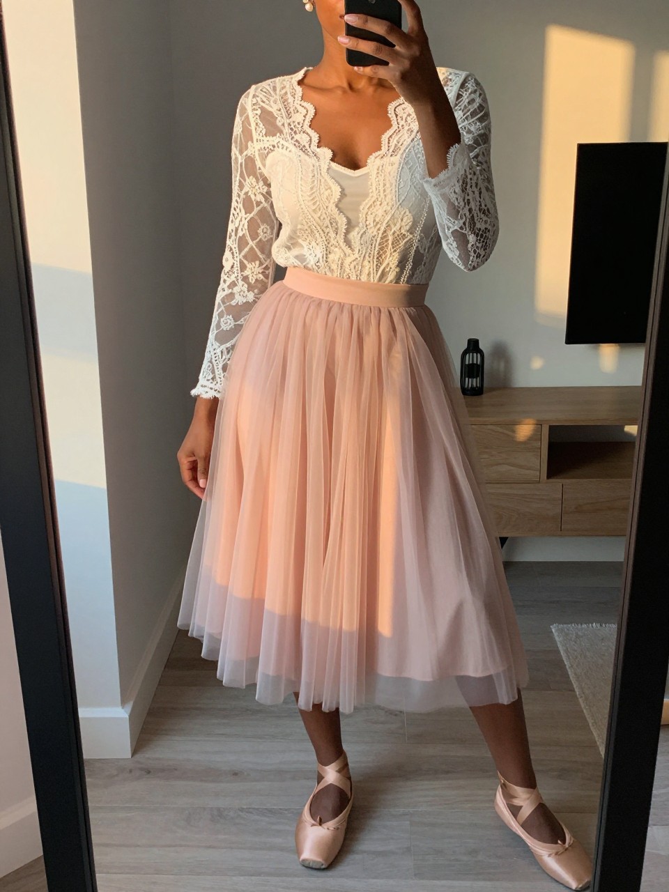 mirror selfie of a black woman wearing a sheer white lace blouse over a white camisole tucked into a blush pink tulle midi skirt with nude ballet flats and pearl stud earrings, standing in a soft romantic pose, cropped at NECK so face doesn't show, in a city apartment, evening golden hour, iPhone photo quality.