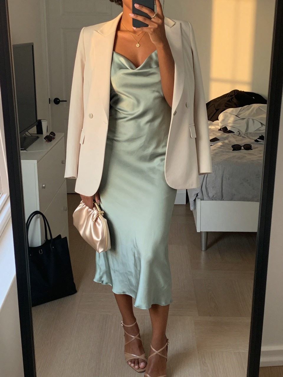 mirror selfie of a black woman wearing a soft sage green satin midi dress with an ivory light blazer, nude leather strappy heels, a champagne satin small clutch, and a delicate gold pendant necklace, holding a phone in front of a mirror, cropped at NECK so face doesn't show, with a natural casual selfie pose, in a city apartment, evening golden hour, including small lived-in details like a tote bag on the floor, sunglasses on a dresser, or soft bedding in the background, latest iPhone photo quality.