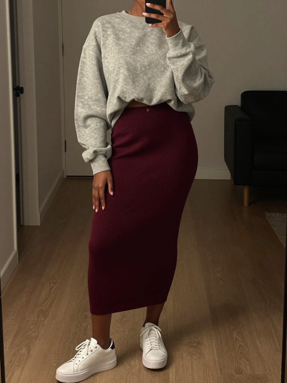 mirror selfie of a black woman wearing an oversized gray sweatshirt half-tucked into a deep burgundy knit midi skirt with white platform sneakers, one hand holding her phone low by her thigh, cropped at NECK so face doesn't show, in a city apartment, warm moody lighting, iPhone photo quality.
