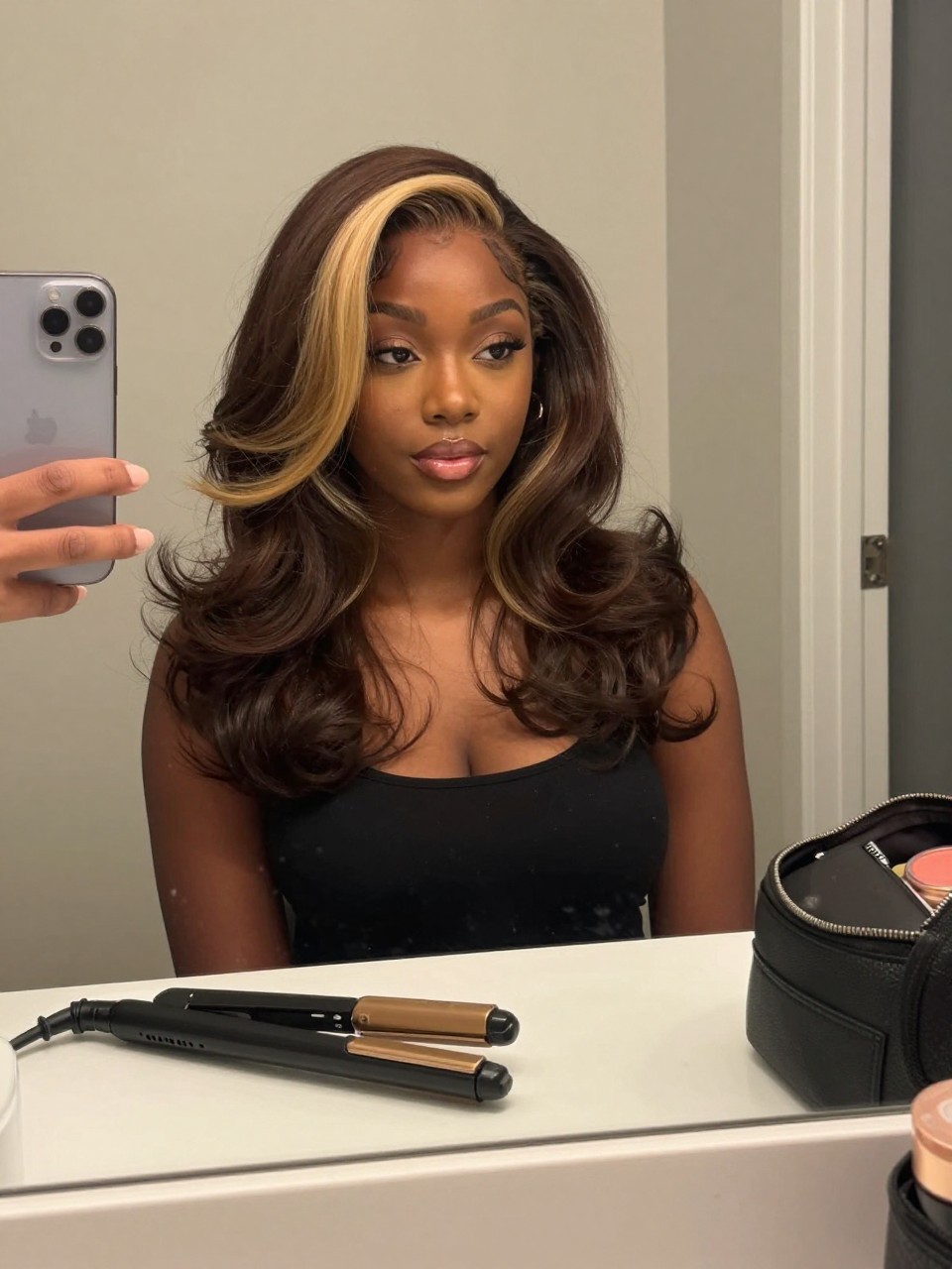 Photo of a black woman wearing brunette hair with a vanilla blonde money piece and soft face-framing waves, candid mirror selfie, bathroom vanity mirror setting, soft indoor lighting, with a flat iron on the counter and a makeup bag nearby, latest iPhone photo quality.