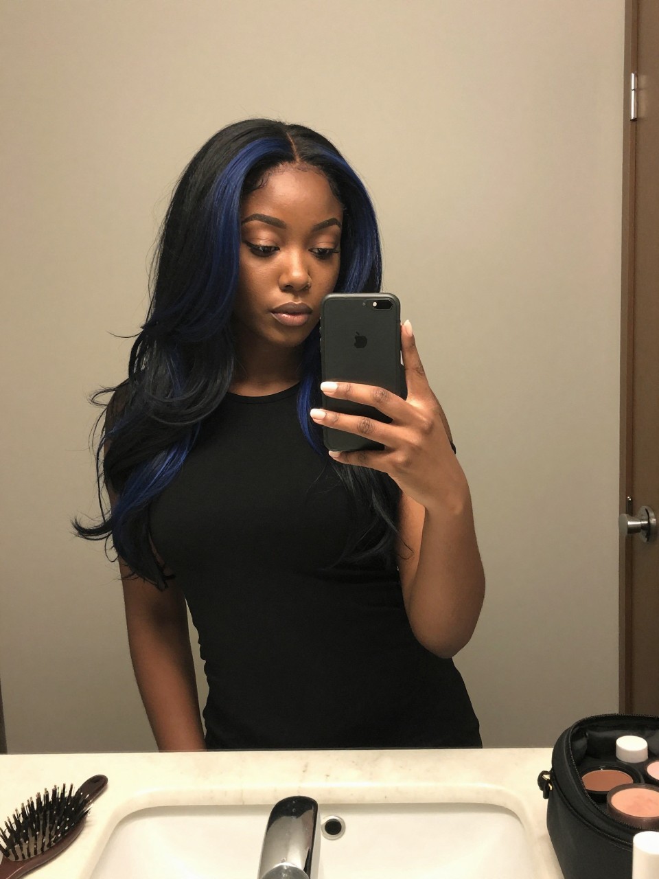 Photo of a black woman wearing a sleek black lob with navy blue face-framing highlights, candid mirror selfie, at a bathroom vanity mirror, soft indoor lighting, containing a small lived-in detail such as a hairbrush on the vanity and a makeup bag nearby, latest iPhone photo quality.