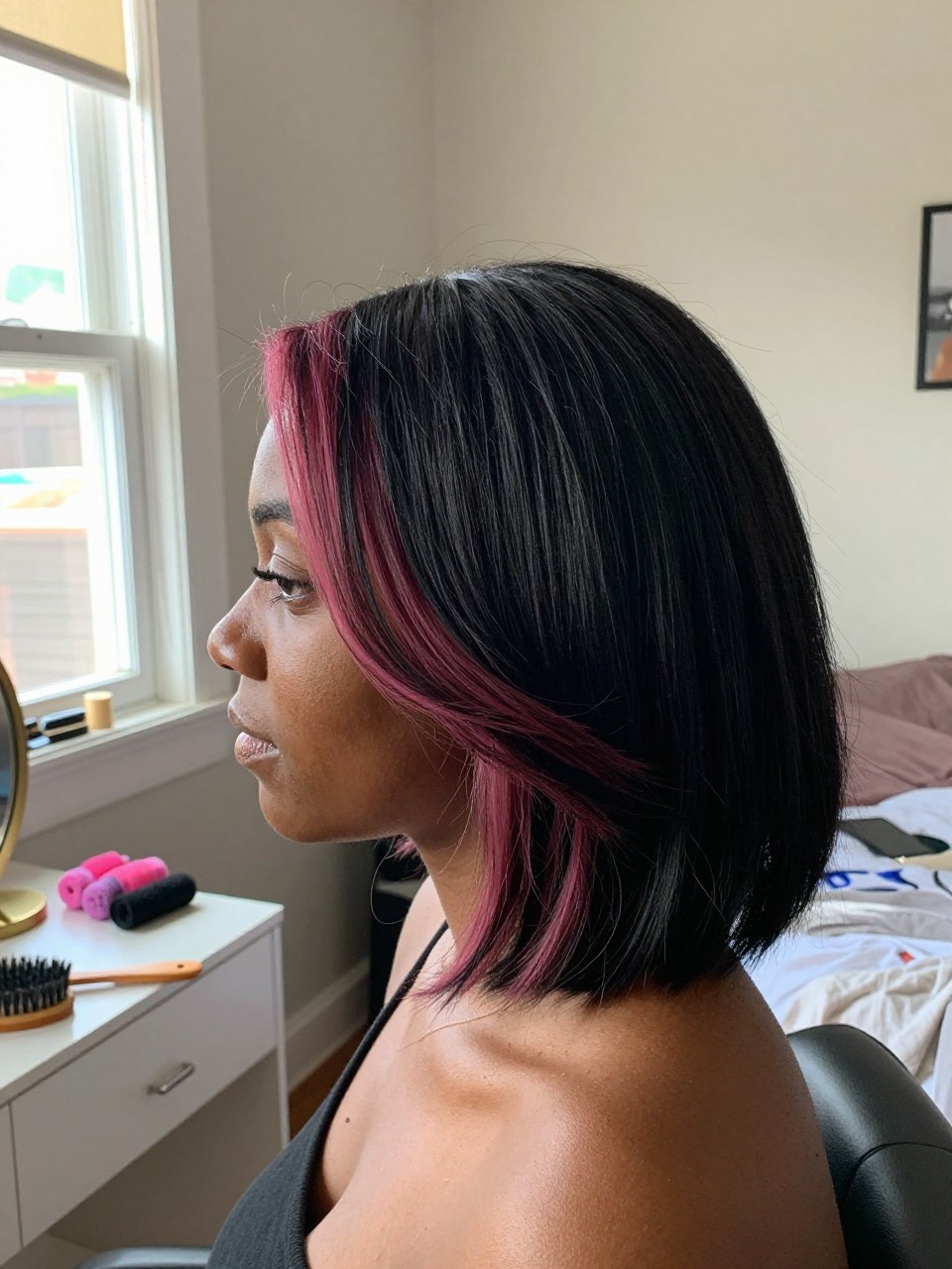 Photo of a black woman wearing a side part black blowout with mauve pink highlights, side profile view, in a sun-drenched bedroom, natural window light, containing a small lived-in detail such as velcro rollers on the vanity and a round brush nearby, latest iPhone photo quality.