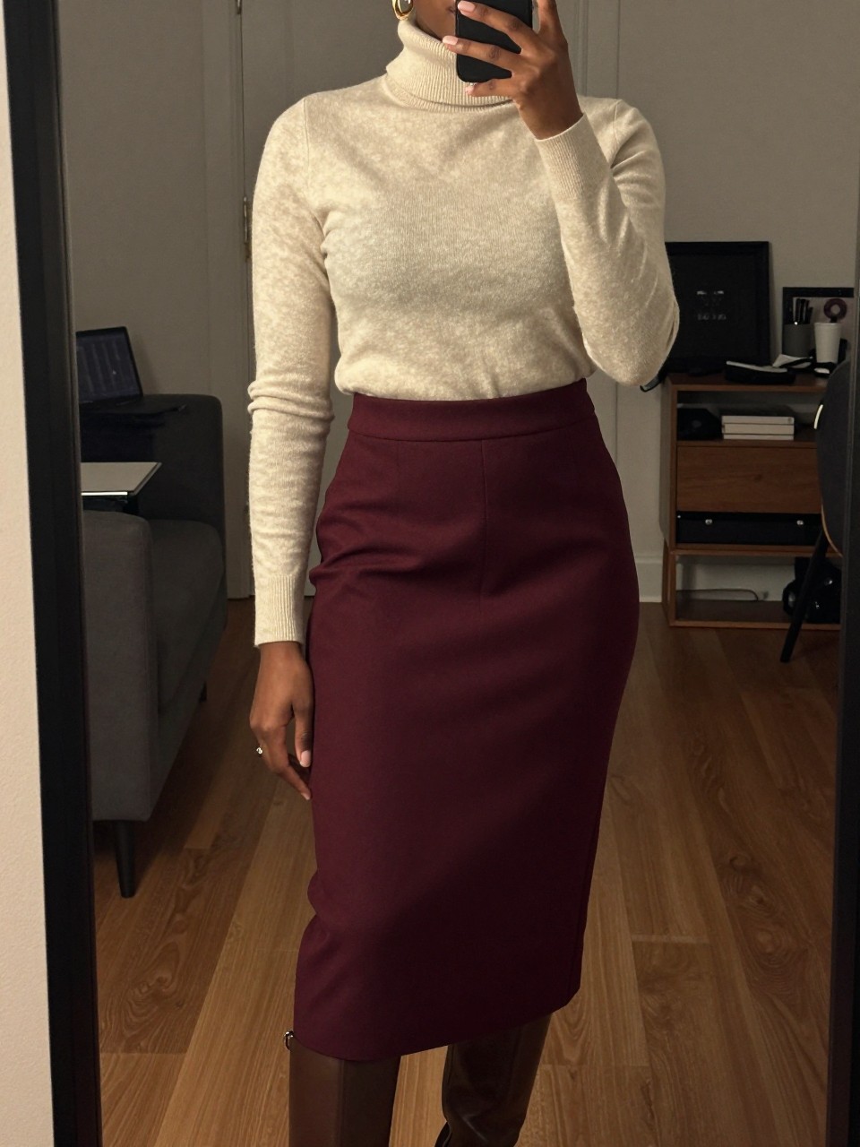 mirror selfie of a black woman wearing a cream turtleneck tucked into a deep burgundy wool midi skirt with gold hoop earrings and brown knee-high boots, one hand adjusting her collar, cropped at NECK so face doesn't show, in a city apartment, warm moody lighting, iPhone photo quality.