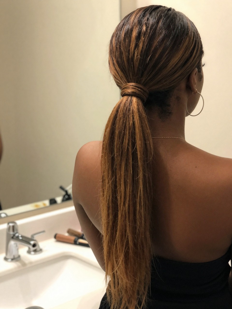 Photo of a Black woman wearing a sleek mid ponytail with dark hair and honey brown highlight panels through the tail, back-of-head view to highlight details, at a bathroom vanity mirror, soft indoor lighting, containing a small lived-in detail such as a flat iron on the counter and hoop earrings peeking through, latest iPhone photo quality.