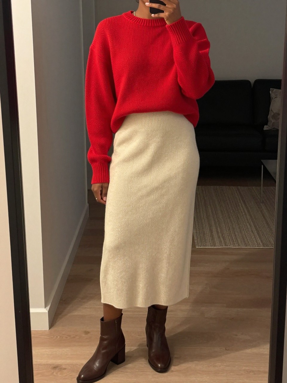 mirror selfie of a black woman wearing a red crewneck sweater tucked into a soft cream knit midi skirt with brown block-heel ankle boots, one hand adjusting her sleeve, cropped at NECK so face doesn't show, in a city apartment, warm moody lighting, iPhone photo quality.