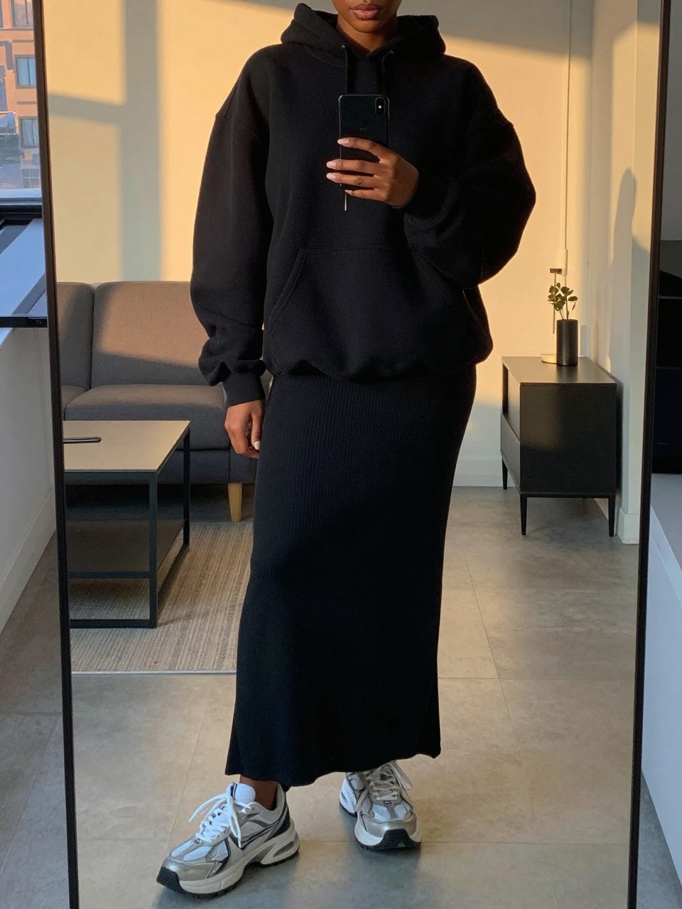 iPhone mirror selfie of a black woman in a black maxi skirt and oversized hoodie with chunky sneakers, standing casually with phone near waist, cropped at NECK so face doesn't show, in a city apartment, evening golden hour, iPhone photo quality.
