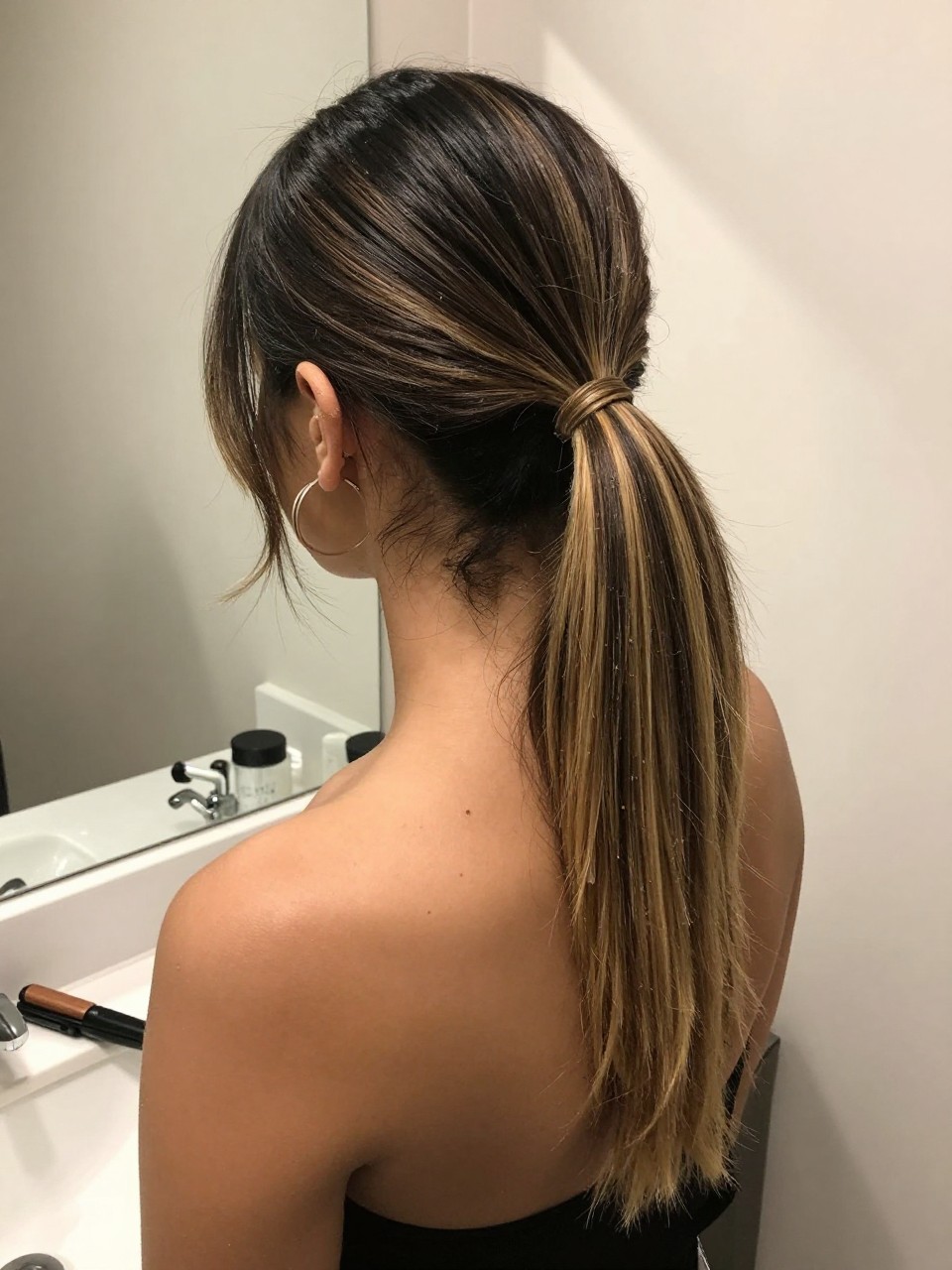 Photo of a woman wearing a sleek mid ponytail with black hair and blonde highlight panels through the tail, back-of-head view to highlight details, at a bathroom vanity mirror, soft indoor lighting, containing a small lived-in detail such as a flat iron on the counter and hoop earrings peeking through, latest iPhone photo quality.