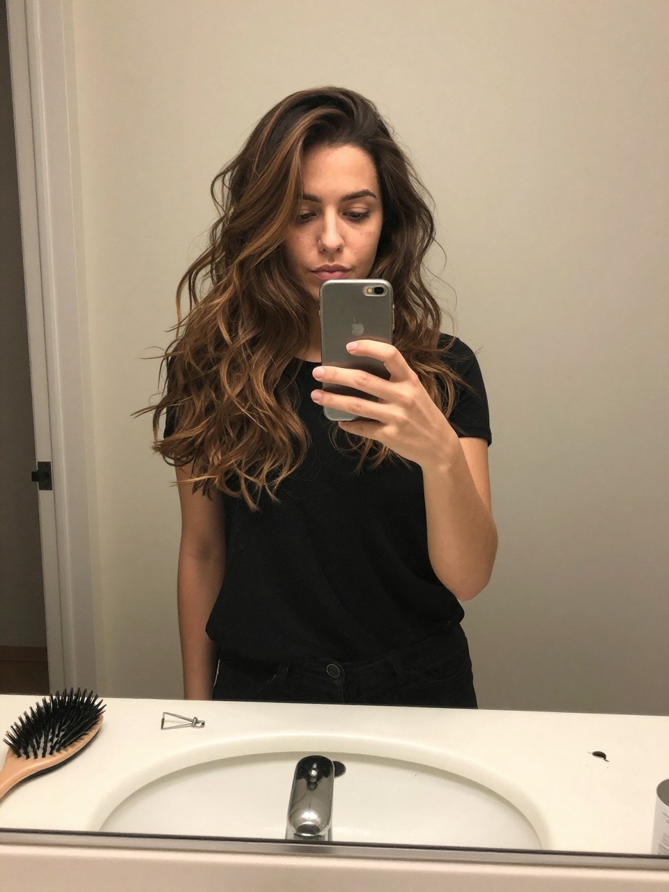 Photo of a woman wearing a wavy brown lob with smoky brunette dark highlights, candid mirror selfie, at a bathroom vanity mirror, soft indoor lighting, containing a small lived-in detail such as a hairbrush on the sink and a silver clip nearby, latest iPhone photo quality.