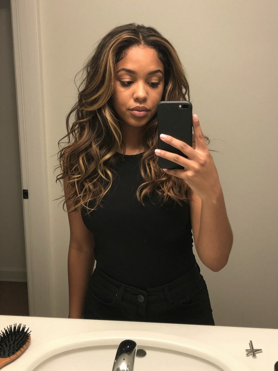 Photo of a biracial woman wearing a wavy dark lob with beige honey highlights, candid mirror selfie, at a bathroom vanity mirror, soft indoor lighting, containing a small lived-in detail such as a hairbrush on the sink and a silver clip nearby, latest iPhone photo quality.