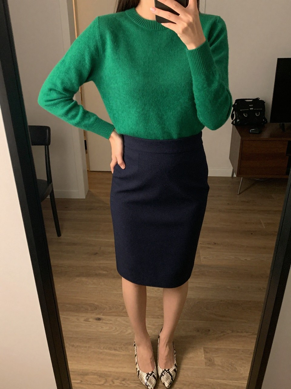 iPhone mirror selfie of a woman wearing an emerald green crewneck sweater tucked into a navy wool pencil skirt with snake print pumps, one hand on her hip, cropped at NECK so face doesn't show, in a city apartment, warm moody lighting, iPhone photo quality.