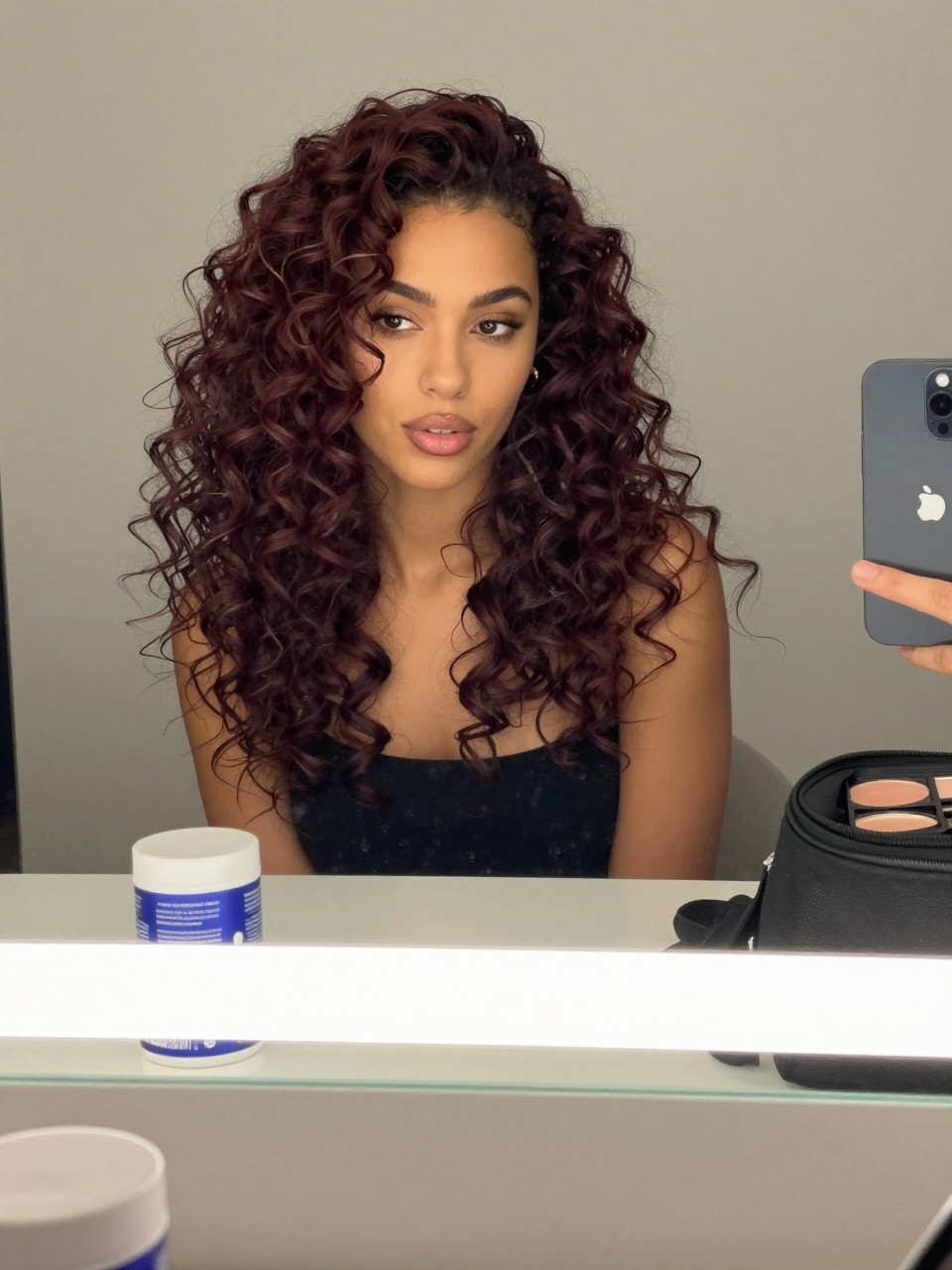 Photo of a woman wearing plum brown curly hair with glossy defined curls, candid mirror selfie, bathroom vanity mirror setting, soft indoor lighting, with a curl cream jar on the counter and a makeup bag nearby, latest iPhone photo quality.