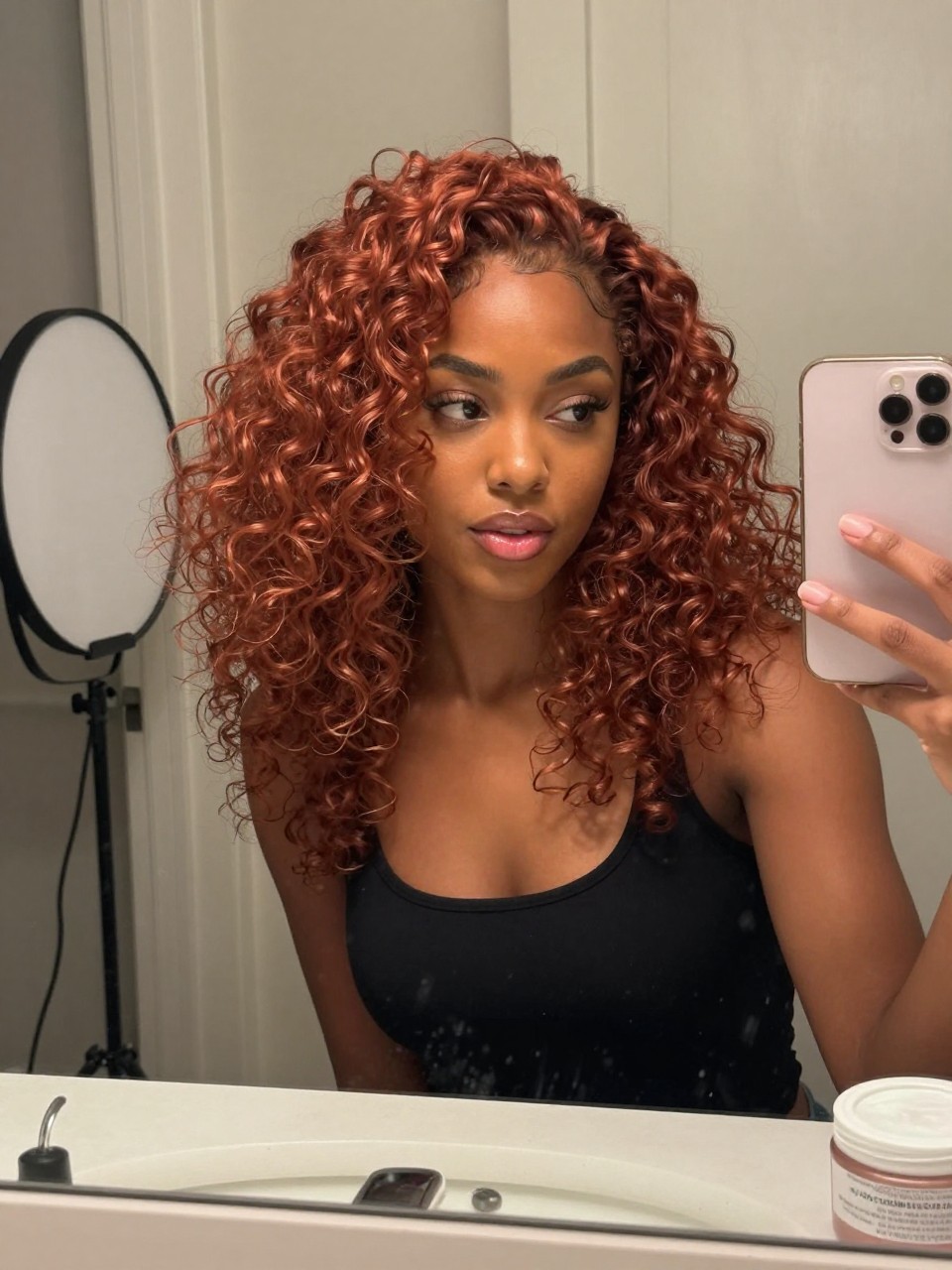 Photo of a black woman wearing strawberry copper curls with soft definition and warm shine, candid mirror selfie, bathroom vanity mirror setting, soft indoor lighting, with a diffuser on the counter and a curl cream jar nearby, latest iPhone photo quality.