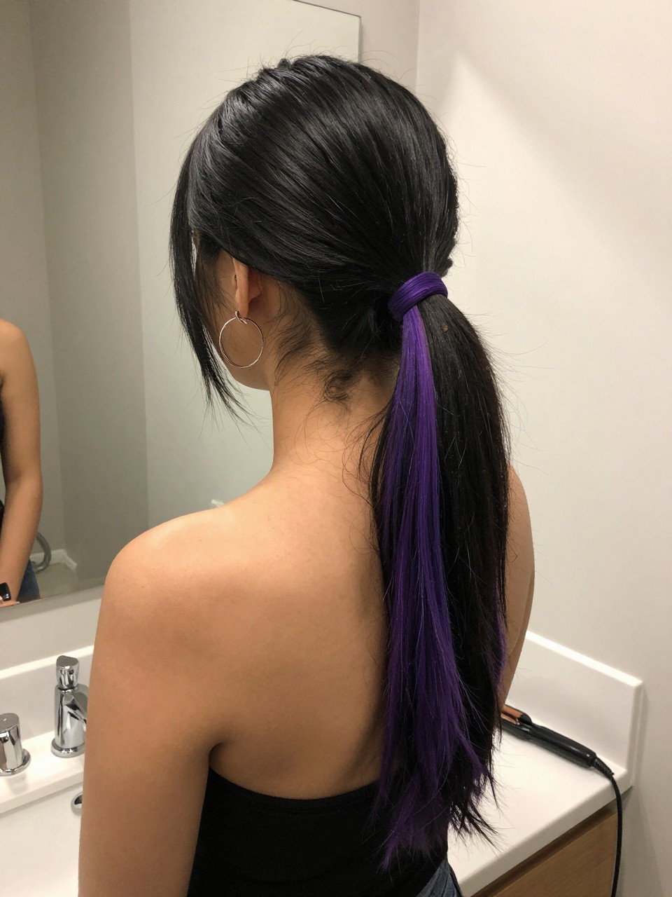 Photo of a woman wearing a sleek mid ponytail with black hair and purple highlight panels through the tail, back-of-head view to highlight details, at a bathroom vanity mirror, soft indoor lighting, containing a small lived-in detail such as a flat iron on the counter and hoop earrings peeking through, latest iPhone photo quality.
