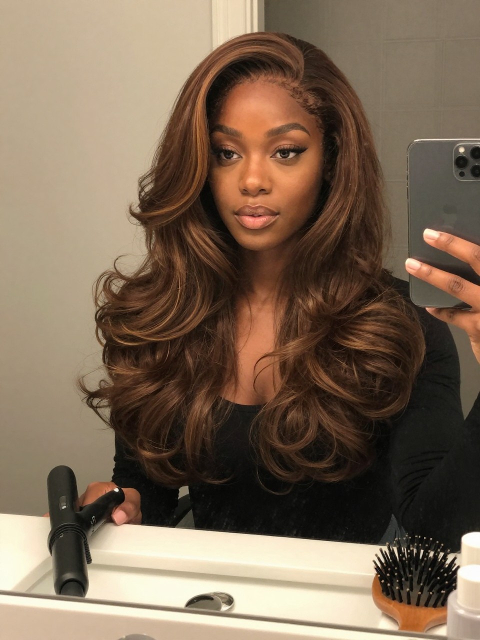 Photo of a black woman wearing chocolate brown hair with honey highlights in soft loose waves, candid mirror selfie, bathroom vanity mirror setting, soft indoor lighting, with a curling wand on the counter and a hairbrush nearby, latest iPhone photo quality.