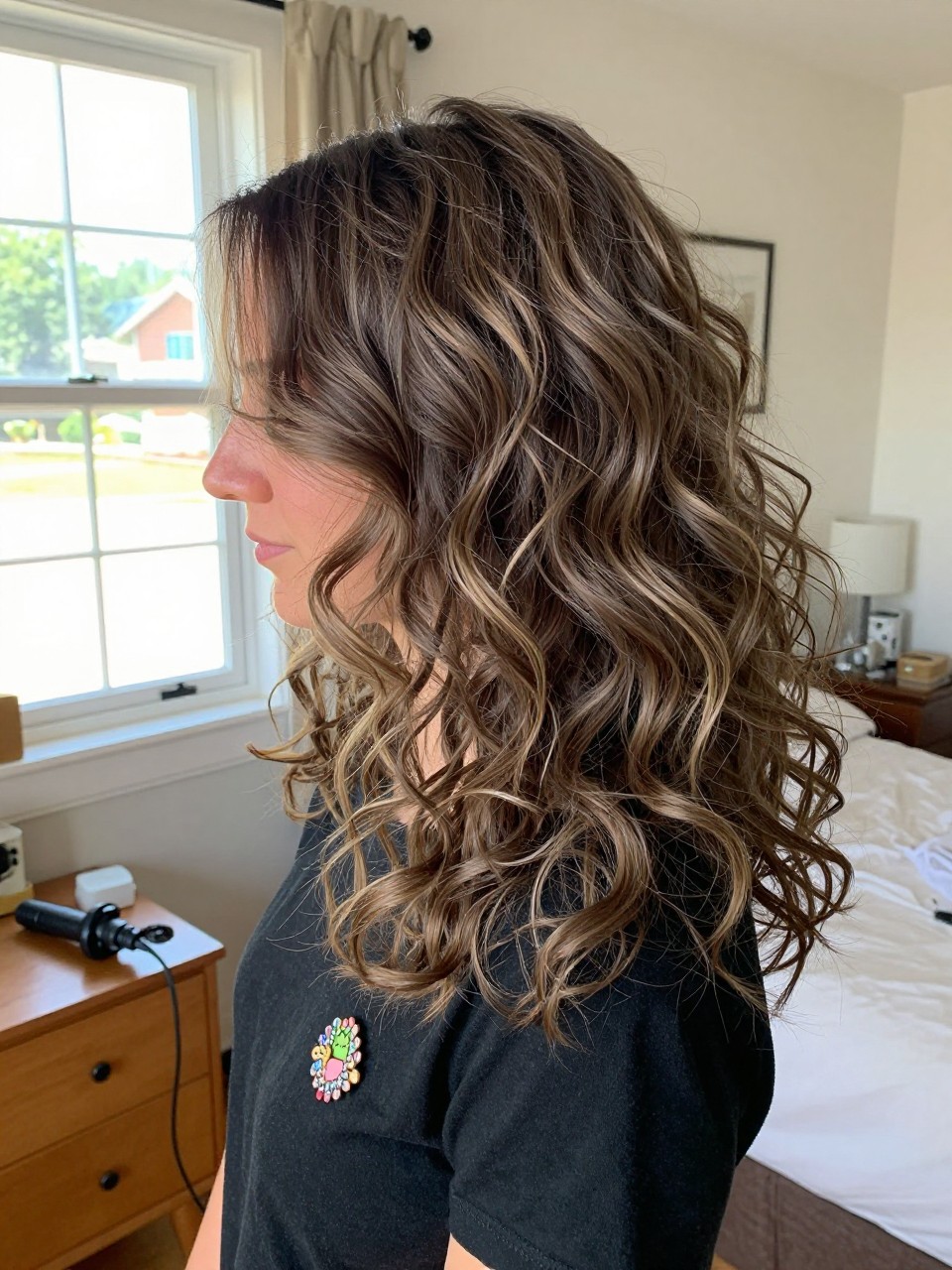 Photo of a woman wearing soft ash brown curls with smoky blonde highlights, side profile view, in a sun-drenched bedroom, natural window light, containing a small lived-in detail such as a decorative pin on one side and a curling iron resting on the nightstand, latest iPhone photo quality.