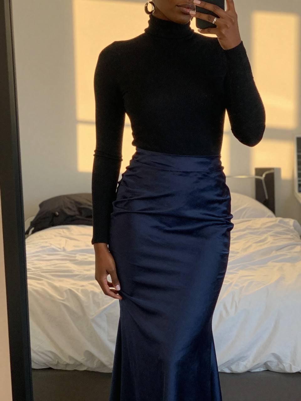 mirror selfie of a black woman in a black turtleneck and a navy satin mermaid skirt with statement earrings, standing with one hand gently touching her earring, cropped at NECK so face doesn't show, in a feminine-themed bedroom, evening golden hour, iPhone photo quality.