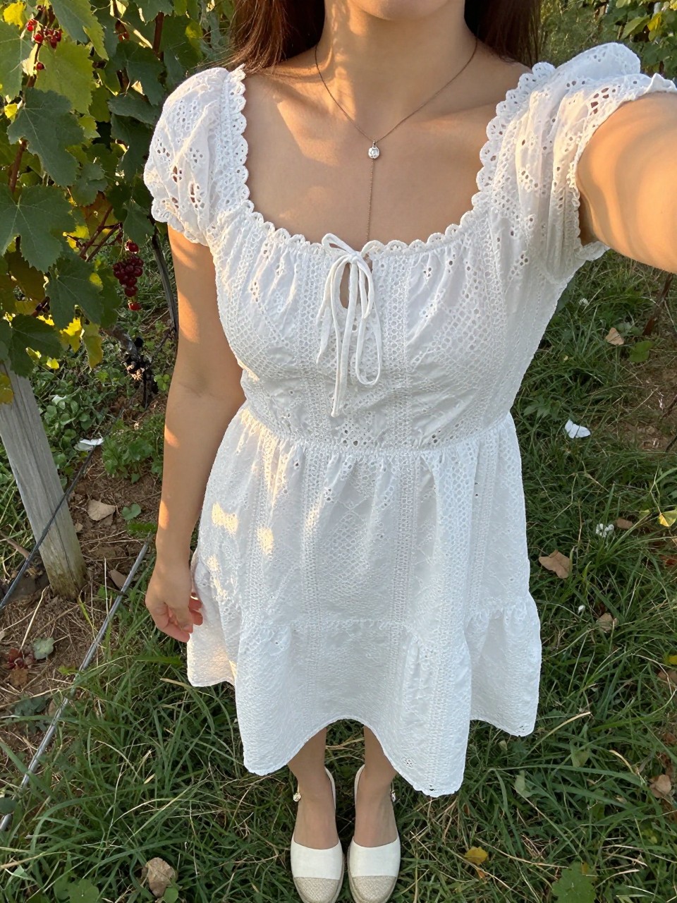 selfie of a woman in a white eyelet dress and espadrilles, standing in a garden or at a winery, cropped at NECK so face doesn't show, at a winery, evening golden hour, iPhone photo quality.