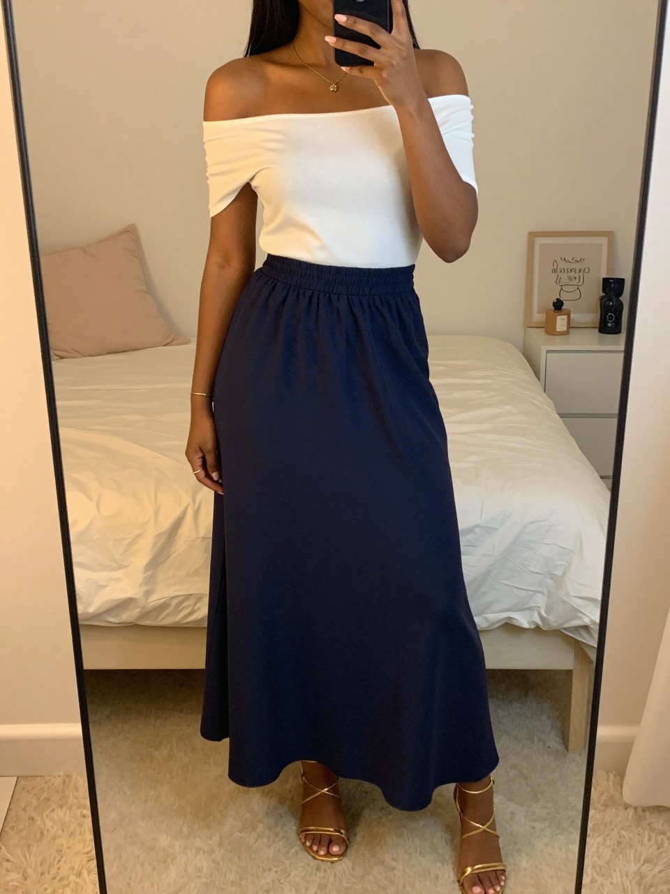 iPhone mirror selfie of a black woman wearing a white off-the-shoulder top tucked into a navy flowy maxi skirt with gold strappy sandals, one hand holding her phone, cropped at NECK so face doesn't show, in a feminine-themed bedroom, evening golden hour, iPhone photo quality.