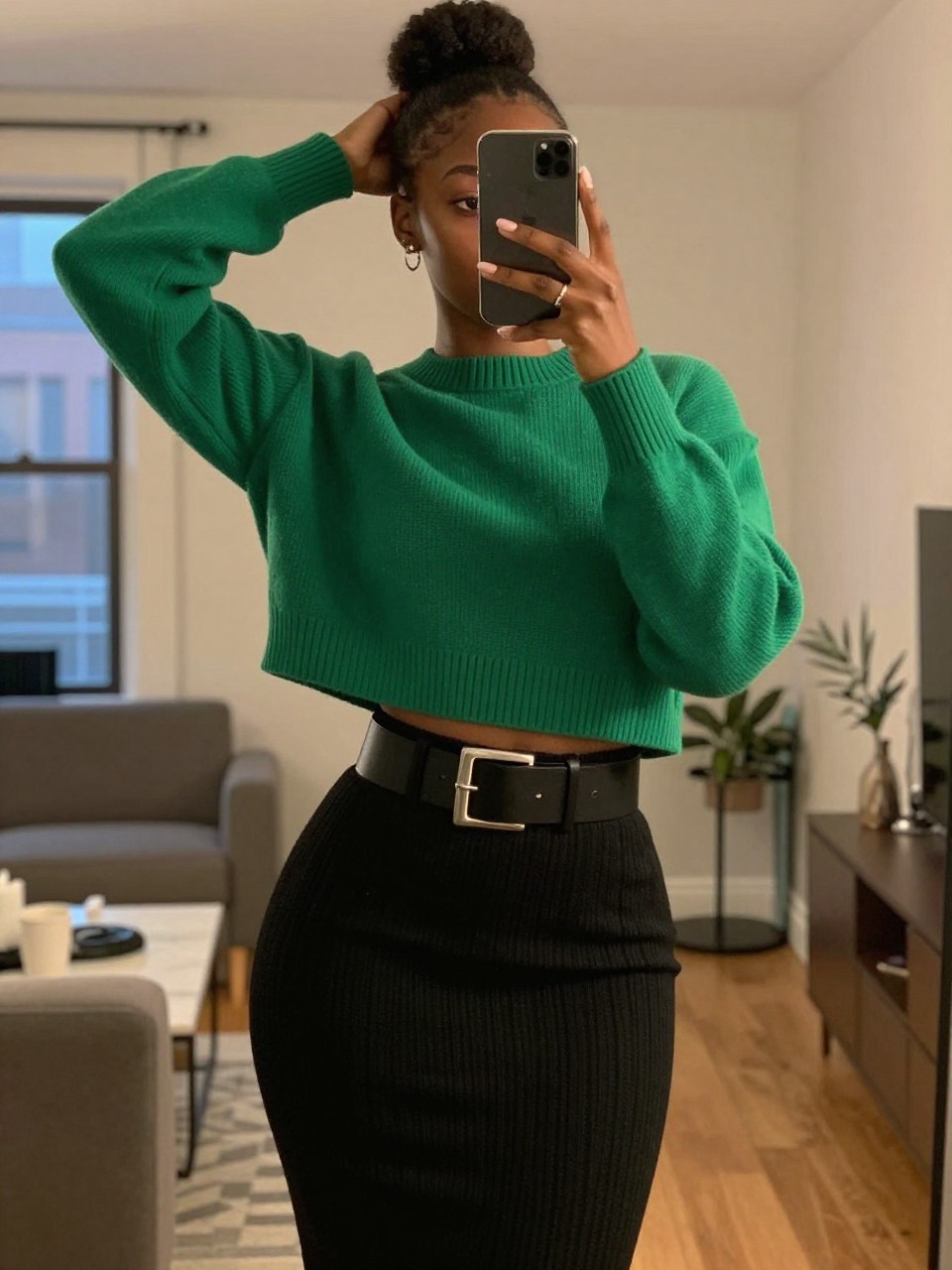 mirror selfie of a black woman wearing an emerald cropped sweater with a black ribbed pencil skirt and a wide silver buckle belt, one hand adjusting her bun, cropped at NECK so face doesn't show, in a city apartment, warm moody lighting, iPhone photo quality.
