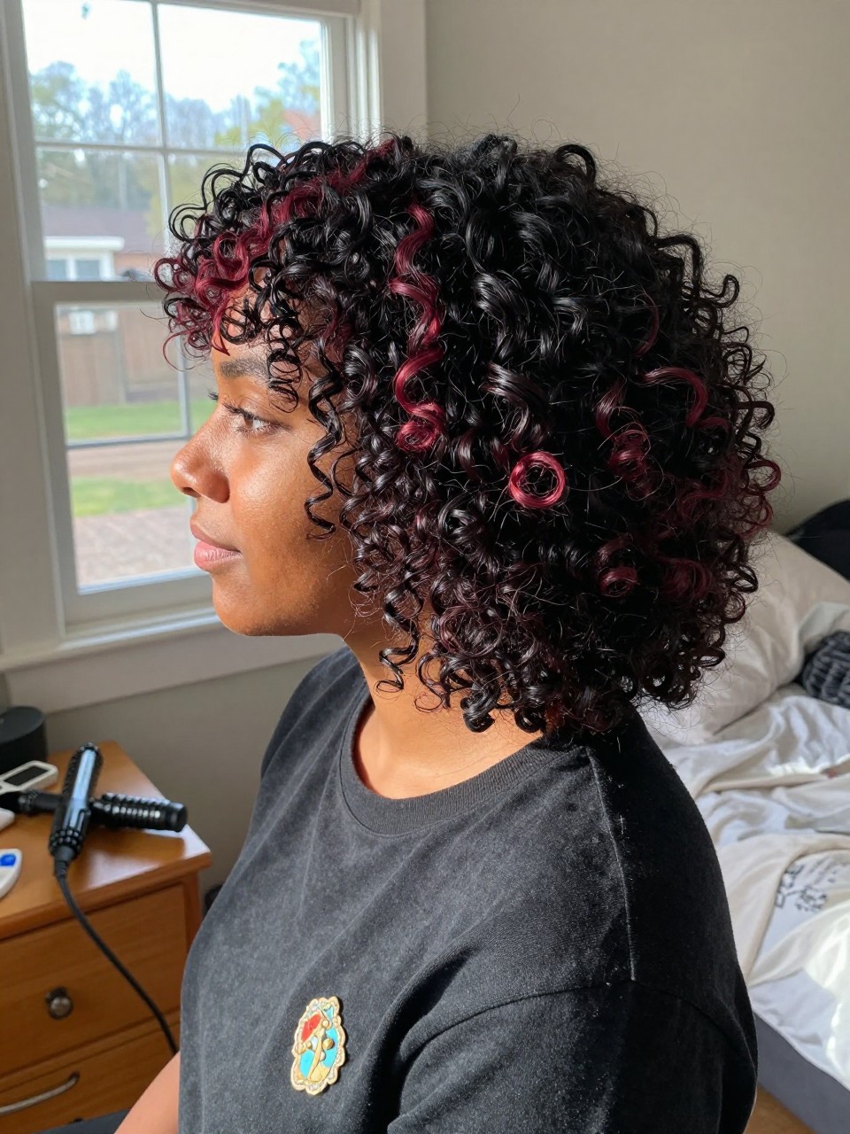Photo of a brown woman wearing soft black curls with garnet red highlight streaks, side profile view, in a sun-drenched bedroom, natural window light, containing a small lived-in detail such as a decorative pin on one side and a curling iron resting on the nightstand, latest iPhone photo quality.