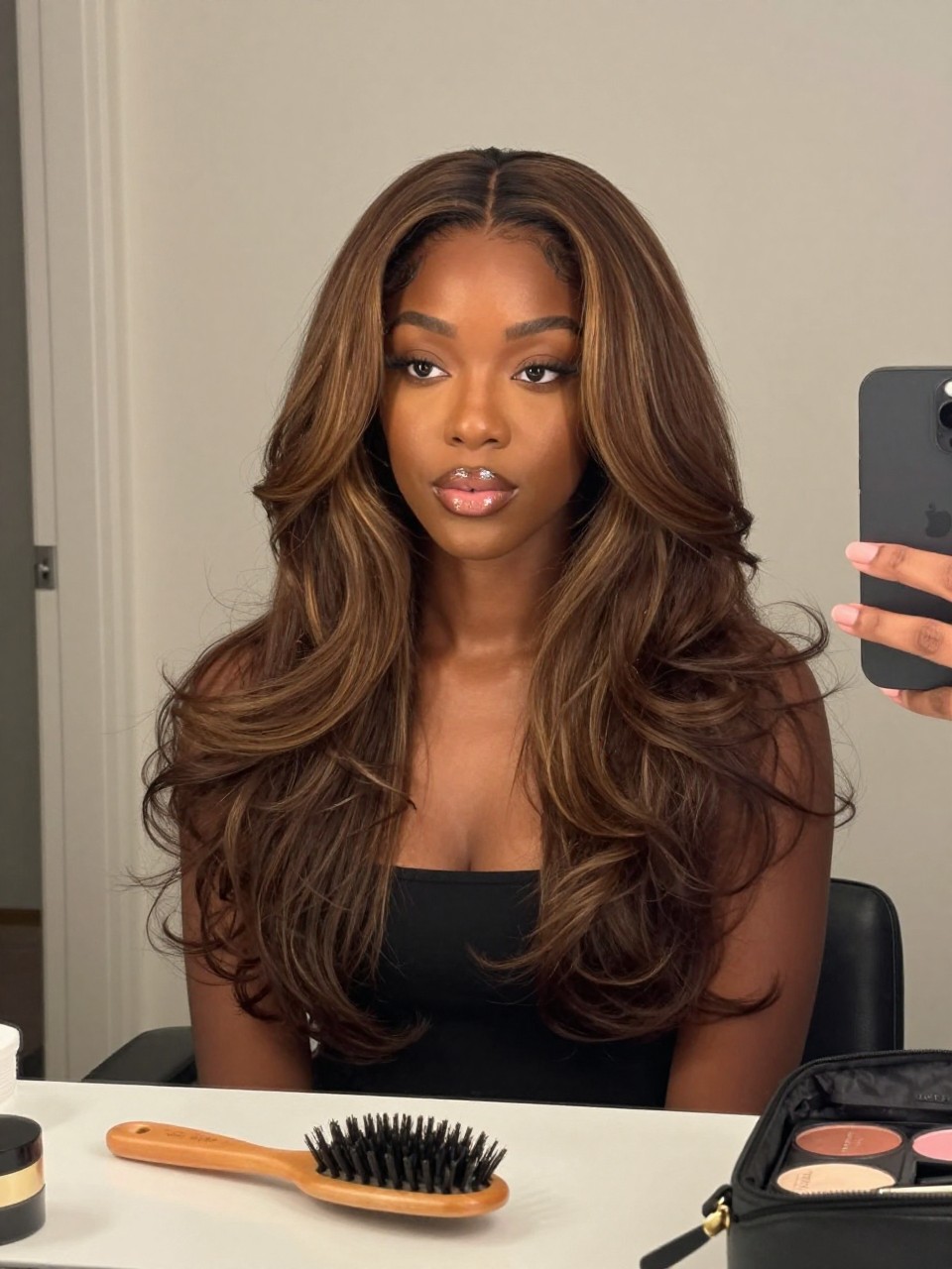 Photo of a black woman wearing brunette hair with soft caramel face-framing highlights and relaxed waves, candid mirror selfie, bathroom vanity mirror setting, soft indoor lighting, with a hairbrush on the vanity and a makeup bag nearby, latest iPhone photo quality.