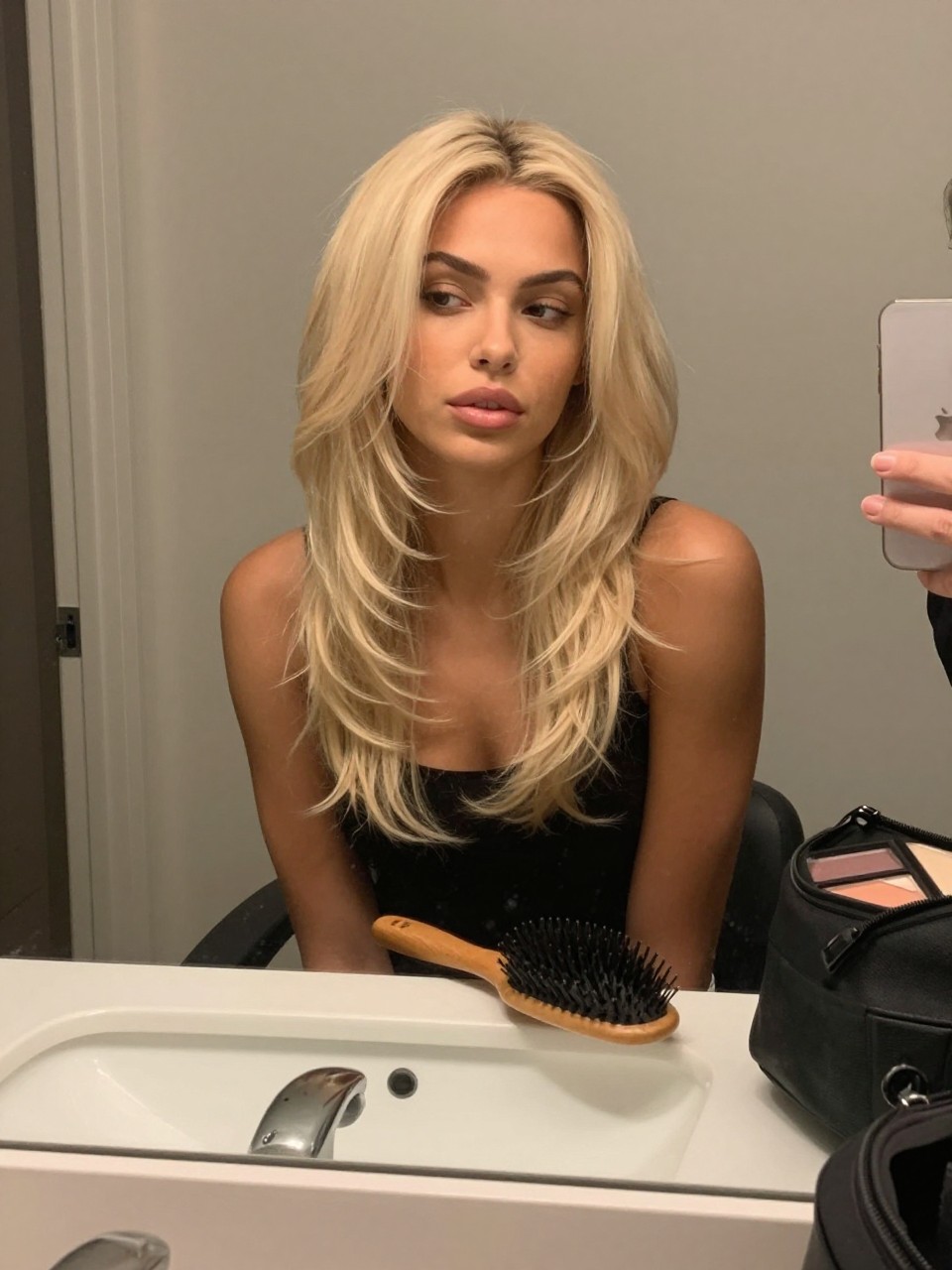 Photo of a biracial woman wearing creamy beige blonde hair with soft face-framing layers, candid mirror selfie, bathroom vanity mirror setting, soft indoor lighting, with a blowout brush on the counter and a makeup bag nearby, latest iPhone photo quality.