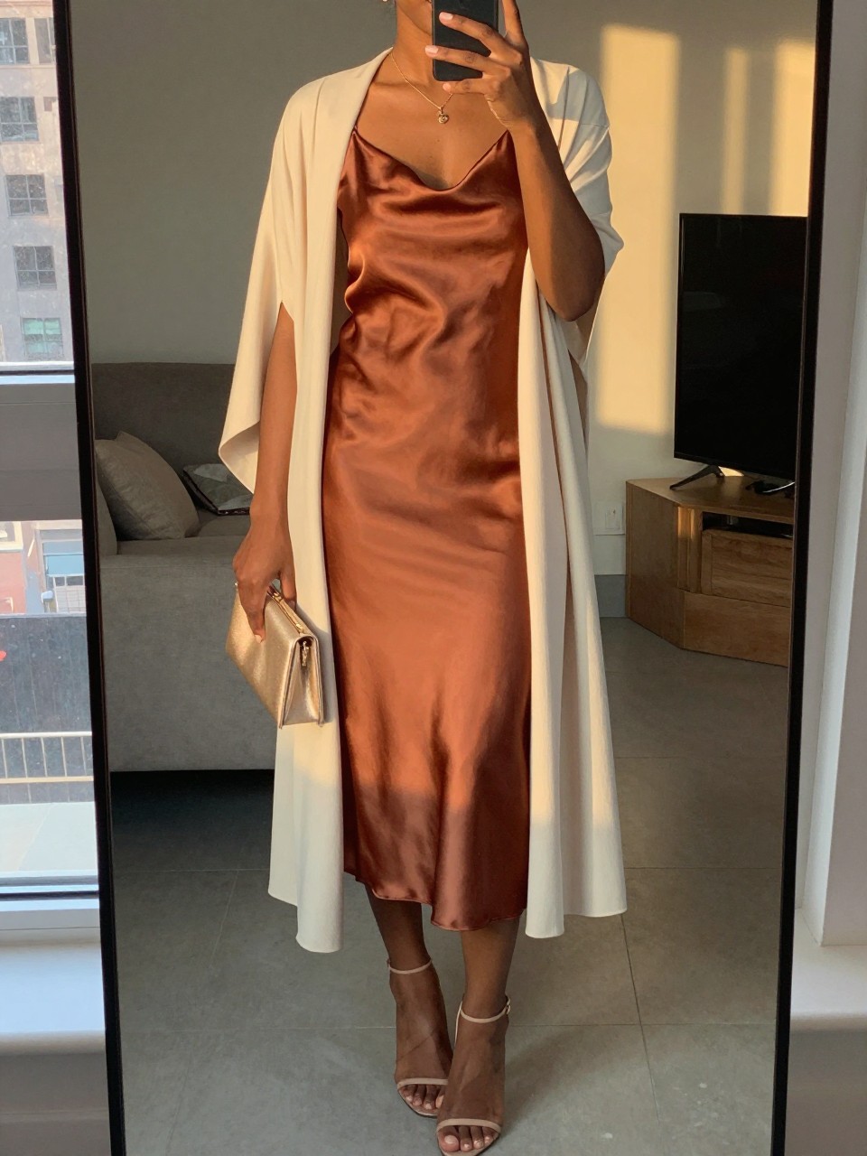 mirror selfie of a black woman wearing a soft copper satin slip midi dress with a cream lightweight duster, nude strappy heels, a delicate gold necklace, and a champagne small clutch, holding a phone in front of a mirror, cropped at NECK so face doesn't show, with a natural casual selfie pose, in a city apartment, evening golden hour, including small lived-in details, latest iPhone photo quality.