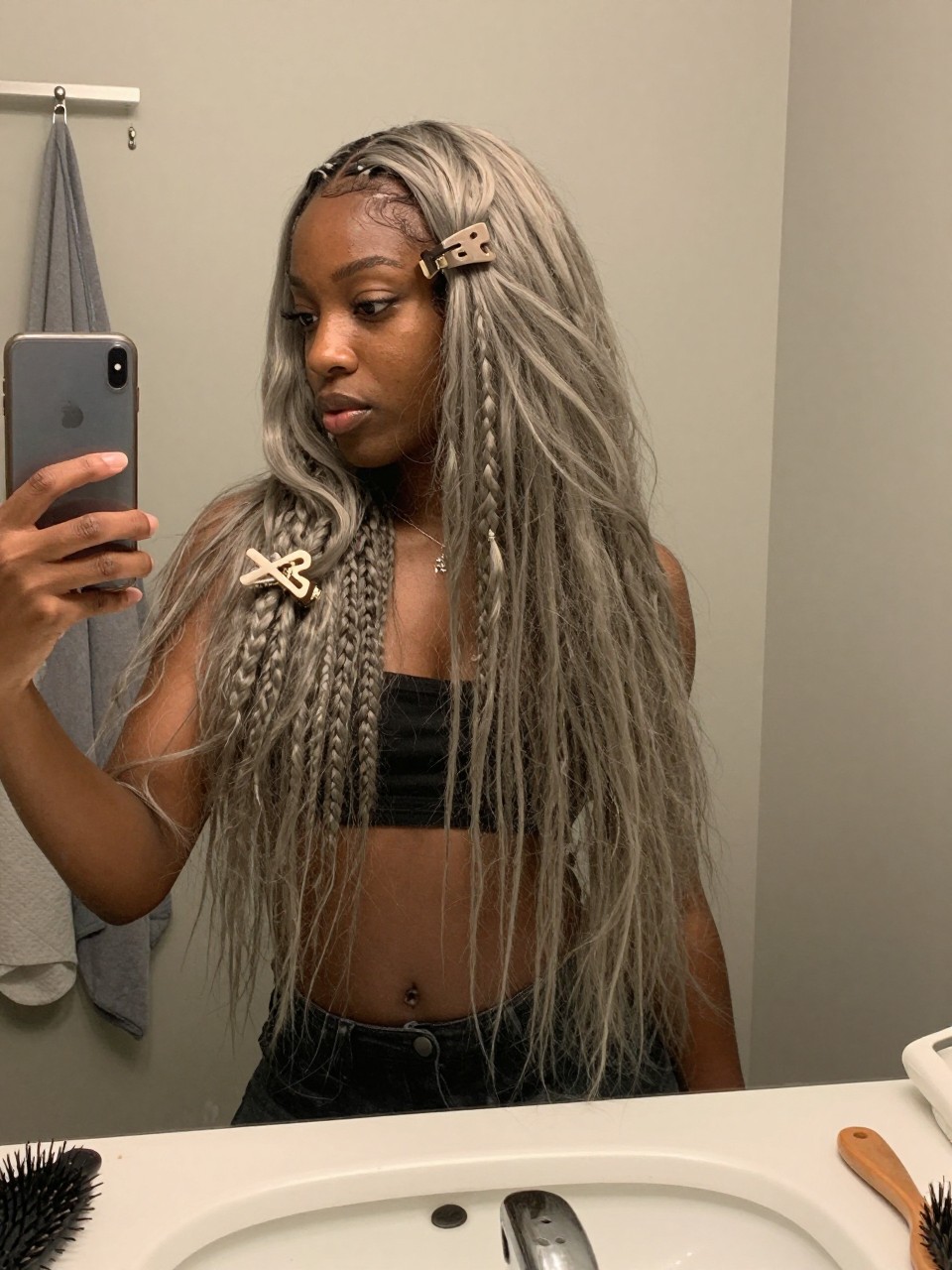 Photo of a black woman wearing a loose claw clip twist with ash beige grey blonde lengths falling softly, candid mirror selfie, at a bathroom vanity mirror, soft indoor lighting, containing a small lived-in detail such as a claw clip clipped to the collar before use and a hairbrush on the sink, latest iPhone photo quality.