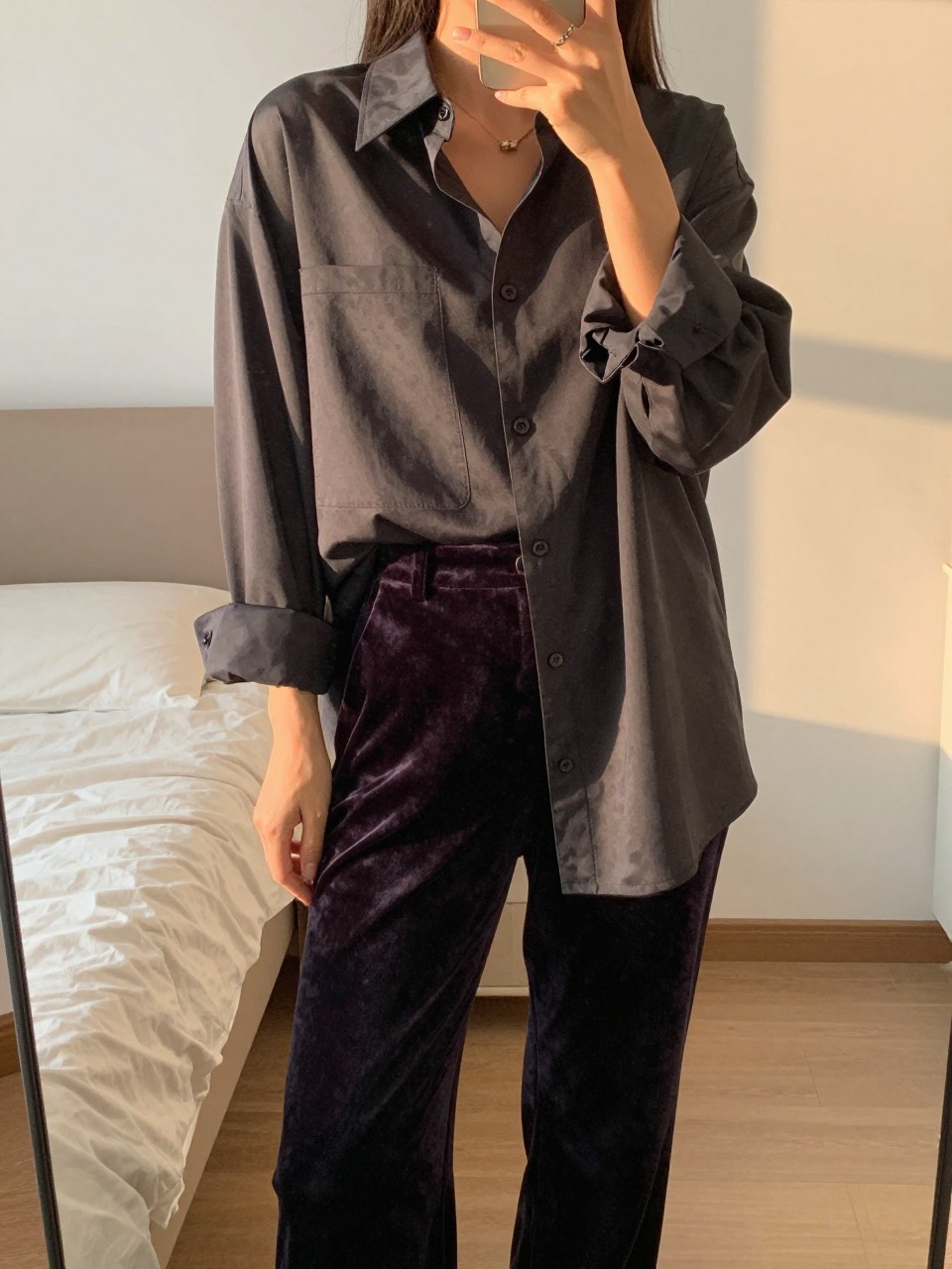 mirror selfie of a woman in an oversized tuxedo shirt and velvet pants, rolling up a sleeve, cropped at NECK so face doesn't show, in a feminine-themed bedroom, evening golden hour, iPhone photo quality.