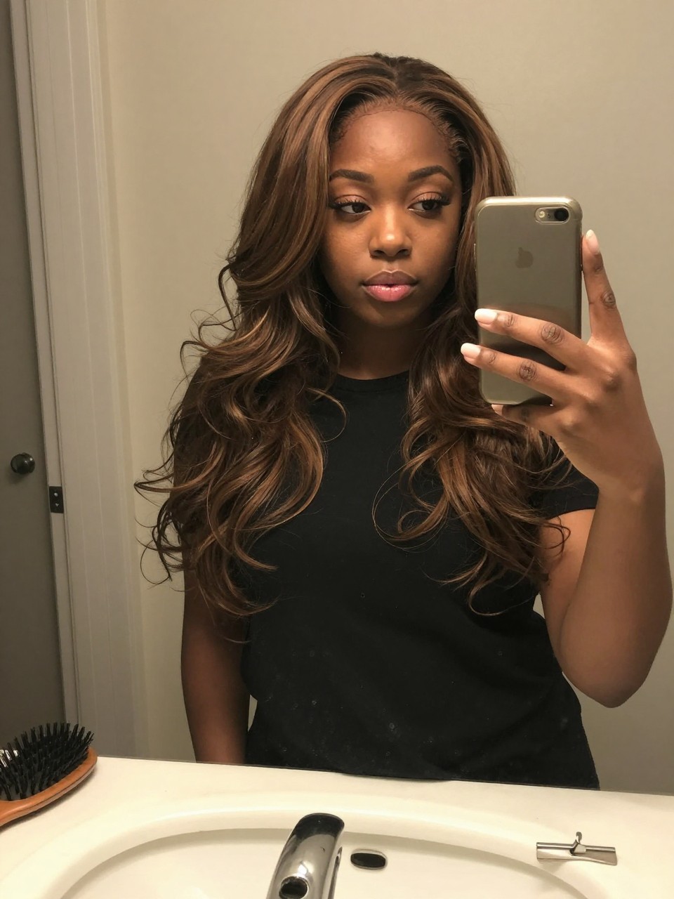 Photo of a black woman wearing a wavy brunette lob with ash blonde highlights, candid mirror selfie, at a bathroom vanity mirror, soft indoor lighting, containing a small lived-in detail such as a hairbrush on the sink and a silver clip nearby, latest iPhone photo quality.