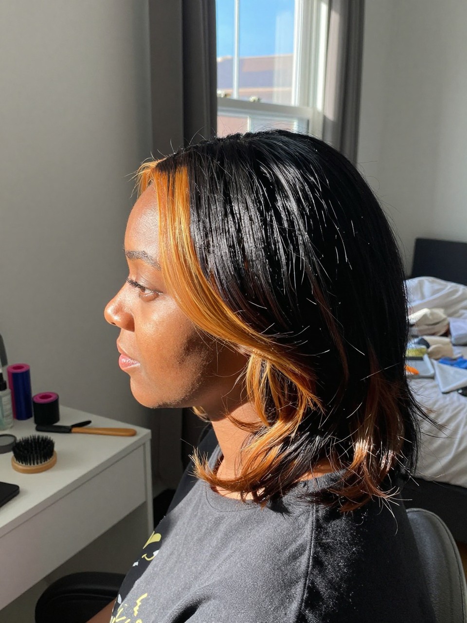 Photo of a black woman wearing a side part black blowout with amber orange highlights, side profile view, in a sun-drenched bedroom, natural window light, containing a small lived-in detail such as velcro rollers on the vanity and a round brush nearby, latest iPhone photo quality.