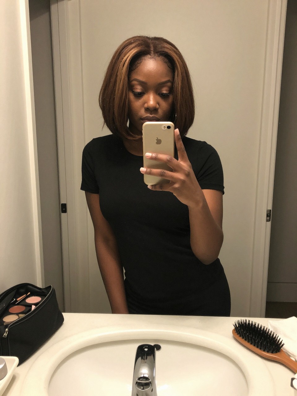 Photo of a black woman wearing a short brown bob with soft highlight money pieces framing the face, candid mirror selfie, at a bathroom vanity mirror, soft indoor lighting, containing a small lived-in detail such as a makeup bag nearby and a hairbrush on the sink, latest iPhone photo quality.