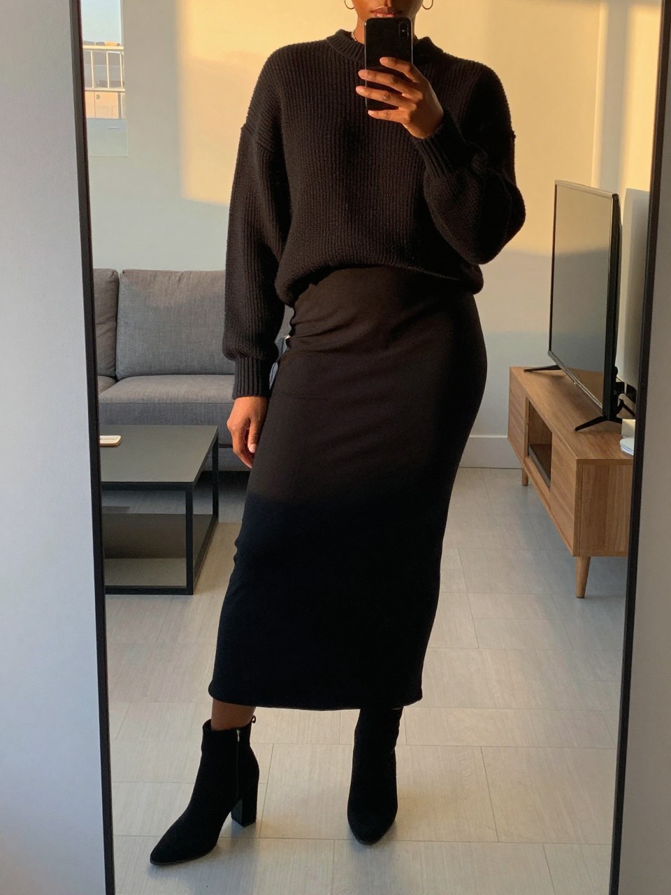 iPhone mirror selfie of a black woman in a black maxi skirt and knit sweater with ankle boots, standing with weight on back leg, cropped at NECK so face doesn't show, in a city apartment, evening golden hour, iPhone photo quality.