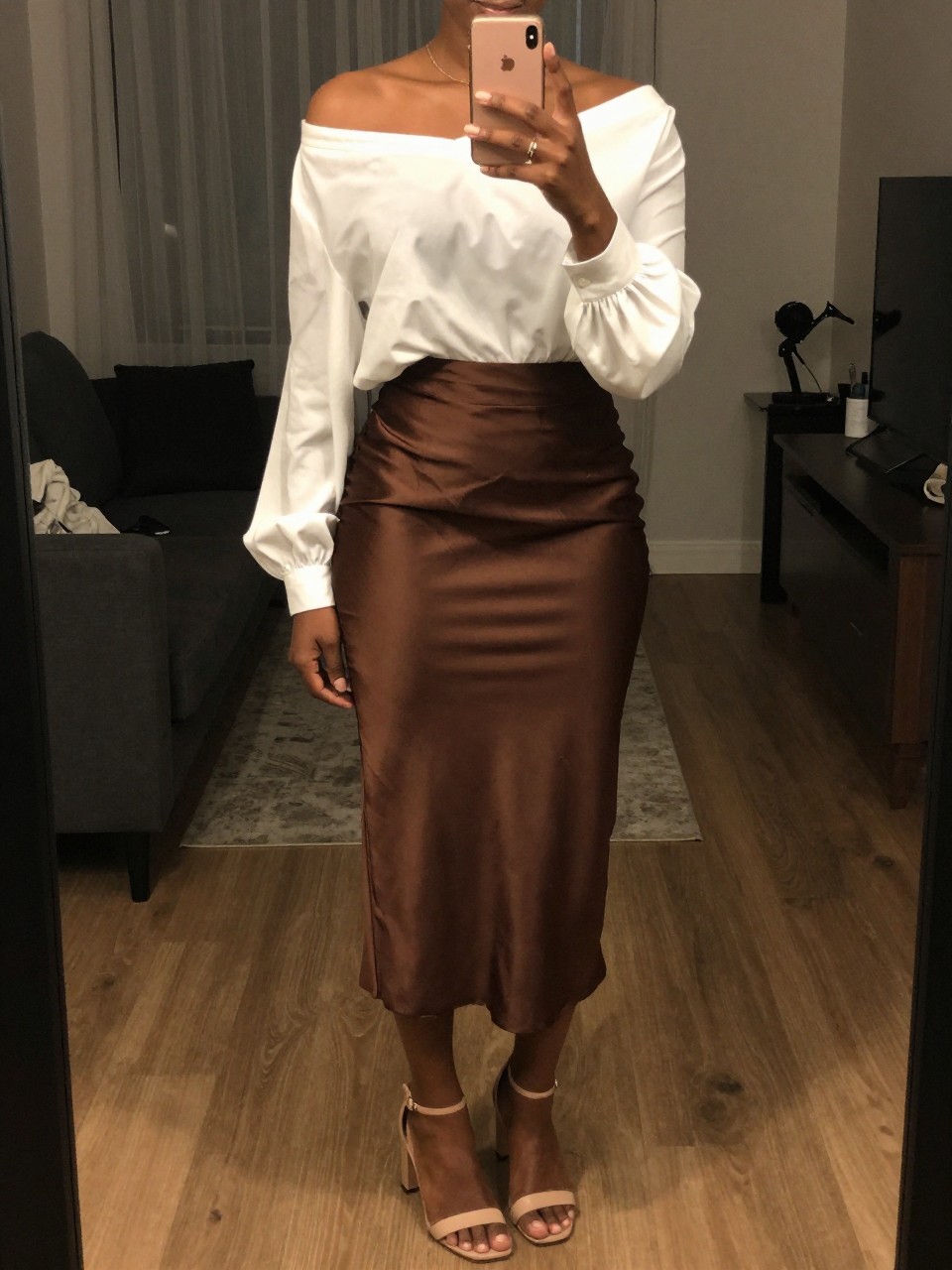 mirror selfie of a black woman wearing a white off-the-shoulder blouse tucked into a chocolate brown satin midi skirt with nude strappy block-heel sandals, one hand holding her phone at chest level, cropped at NECK so face doesn't show, in a city apartment, warm moody lighting, iPhone photo quality.