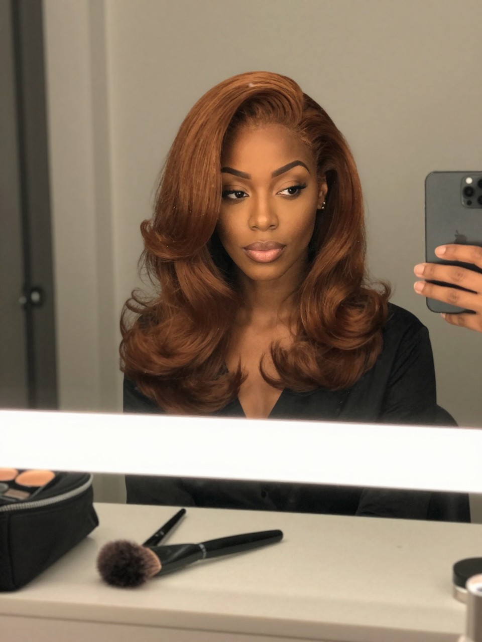 Photo of a black woman wearing soft copper brown brunette hair in a smooth bouncy blowout, candid mirror selfie, bathroom vanity mirror setting, soft indoor lighting, with a round brush on the counter and a makeup bag nearby, latest iPhone photo quality.