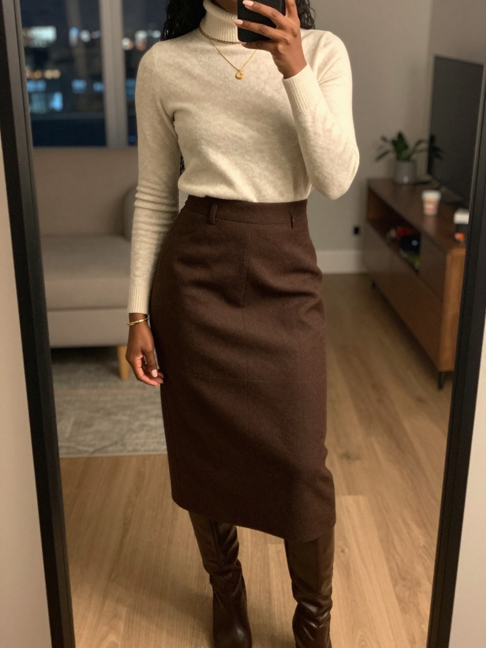 iPhone mirror selfie of a black woman wearing a cream turtleneck tucked into a rich chocolate brown wool midi skirt with gold jewelry and brown knee-high boots, one hand adjusting her collar, cropped at NECK so face doesn't show, in a city apartment, warm moody lighting, iPhone photo quality.
