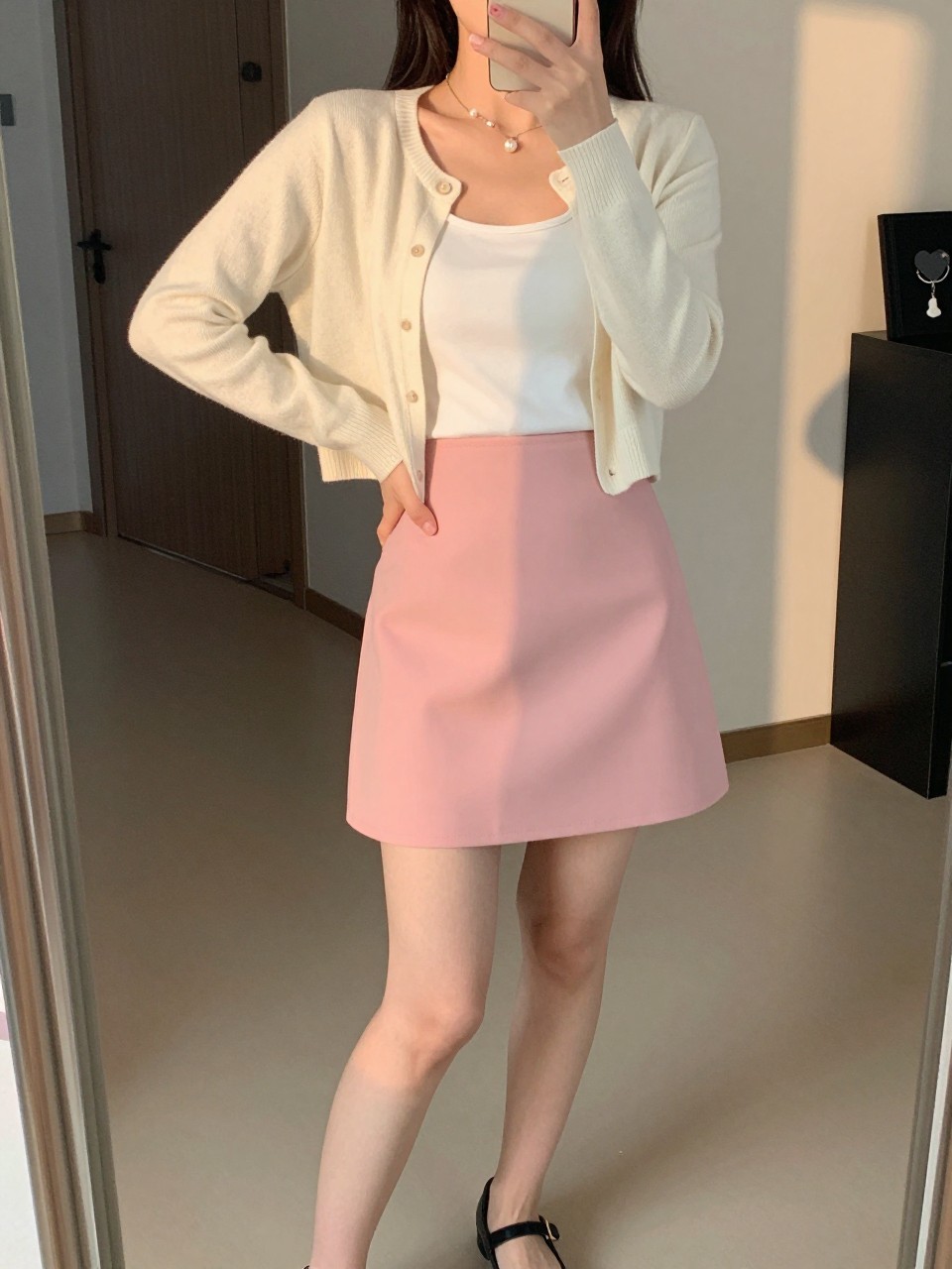 mirror selfie of a woman wearing a cream cropped cardigan over a white tank top tucked into a bubblegum pink A-line mini skirt with black Mary Jane flats and pearl stud earrings, standing in a sweet feminine pose, cropped at NECK so face doesn't show, in a city apartment, evening golden hour, iPhone photo quality.