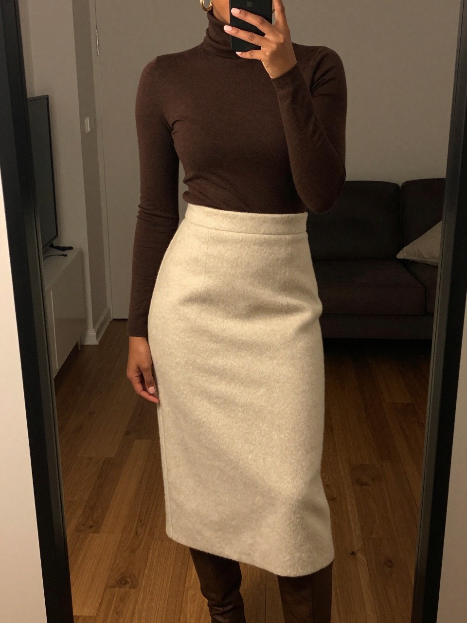 iPhone mirror selfie of a black woman wearing a chocolate brown turtleneck tucked into a soft cream wool midi skirt with gold hoop earrings and brown knee-high boots, one hand adjusting her collar, cropped at NECK so face doesn't show, in a city apartment, warm moody lighting, iPhone photo quality.
