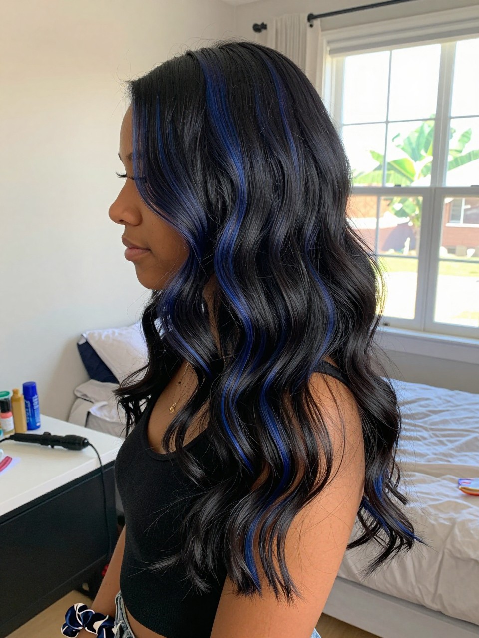Photo of a biracial woman wearing soft black waves with midnight blue highlight ribbons woven through the lengths, side profile view, in a sun-drenched bedroom, natural window light, containing a small lived-in detail such as a curling iron on the counter and a silk scrunchie on the wrist, latest iPhone photo quality.
