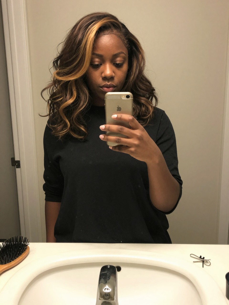 Photo of a Black woman wearing a wavy dark lob with beige honey brown highlights, candid mirror selfie, at a bathroom vanity mirror, soft indoor lighting, containing a small lived-in detail such as a hairbrush on the sink and a silver clip nearby, latest iPhone photo quality.