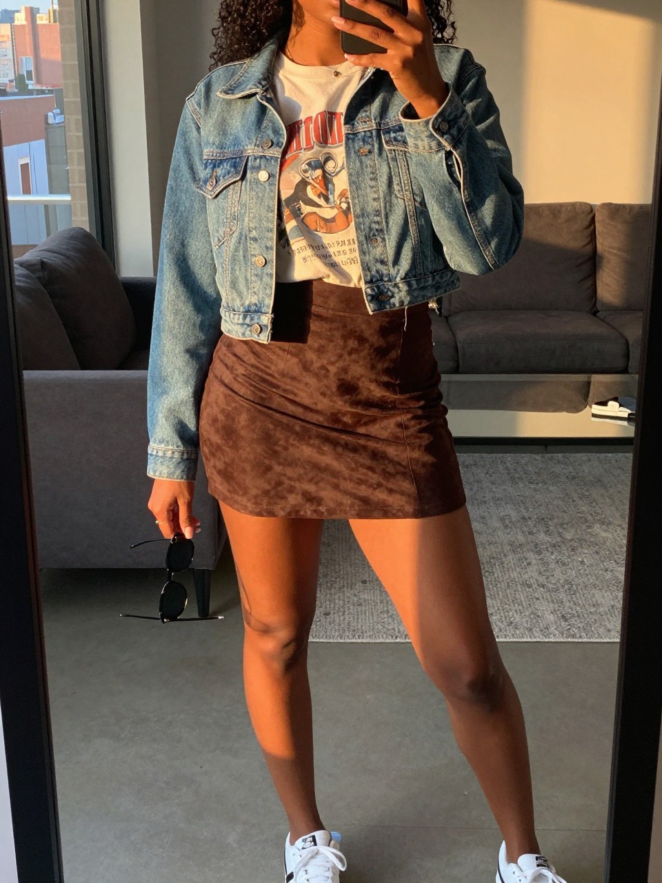 mirror selfie of a black woman wearing a vintage band tee tucked into a chocolate brown suede mini skirt with a cropped light-wash denim jacket and white sneakers, holding sunglasses in one hand, standing in a casual pose, cropped at NECK so face doesn't show, in a city apartment, evening golden hour, iPhone photo quality.