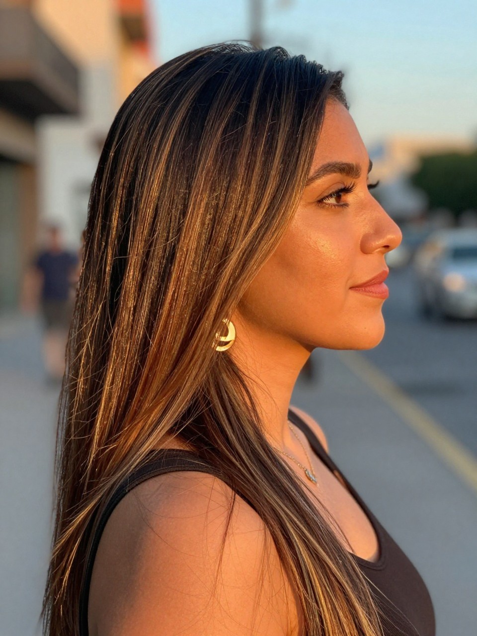 Photo of a mexican woman wearing long straight dark hair with honey face-framing highlights, side profile view, in a softly blurred city street background, golden hour glow, containing a small lived-in detail such as statement earrings peeking through and a crossbody strap on the shoulder, latest iPhone photo quality.