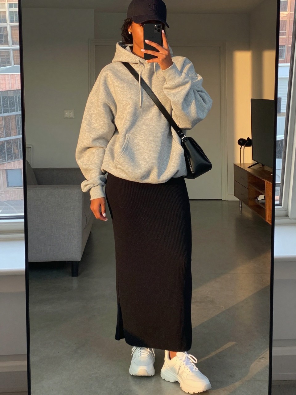 mirror selfie of a black woman wearing an oversized heather grey hoodie that covers most of a black jersey knit maxi skirt with chunky white sneakers and a baseball cap, holding a black crossbody bag, standing in a relaxed casual pose, cropped at NECK so face doesn't show, in a city apartment, evening golden hour, iPhone photo quality.