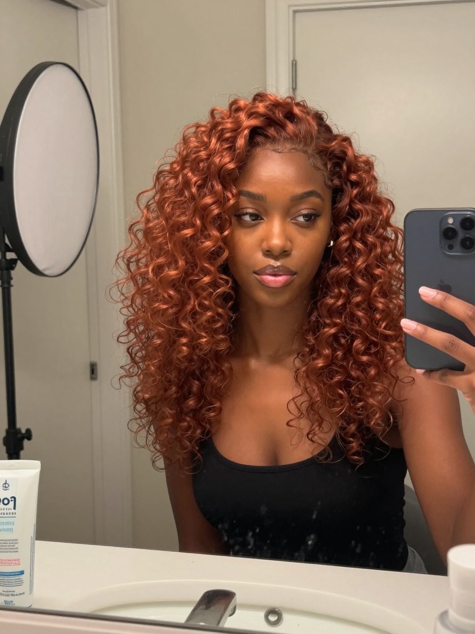 Photo of a black woman wearing strawberry copper curls with soft definition and warm shine, candid mirror selfie, bathroom vanity mirror setting, soft indoor lighting, with a diffuser on the counter and a curl cream jar nearby, latest iPhone photo quality.