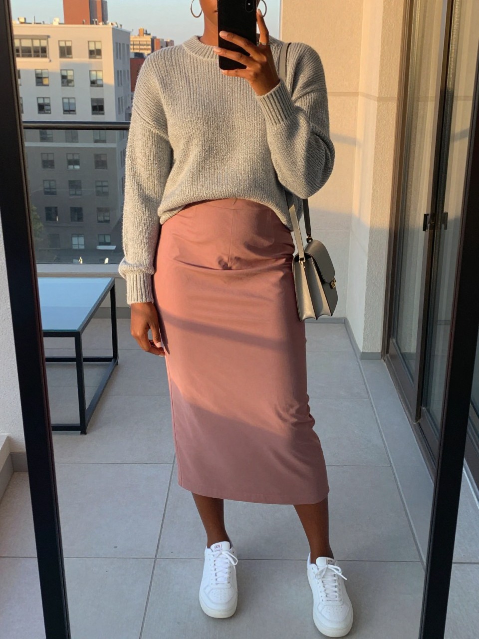iPhone mirror selfie of a black woman wearing a heather grey fine-knit sweater tucked into a dusty rose midi skirt with white sneakers and silver hoop earrings, holding a grey crossbody bag, standing in an elegant relaxed pose, cropped at NECK so face doesn't show, in a city apartment, evening golden hour, iPhone photo quality.