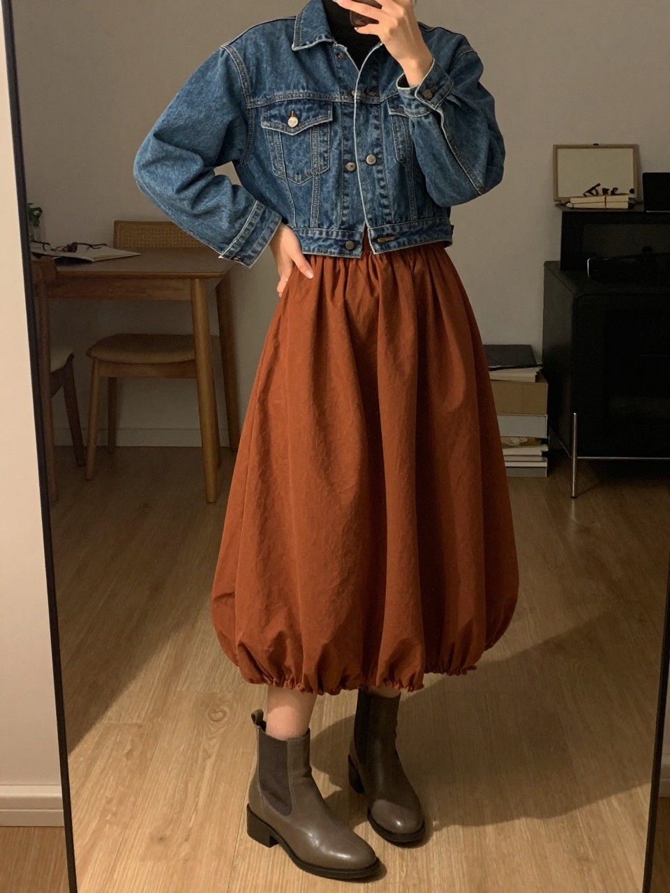 mirror selfie of a woman in a cropped denim jacket and a rust-colored balloon skirt with ankle boots, one hand resting on her hip, cropped at NECK so face doesn't show, in a city apartment, warm moody lighting, iPhone photo quality.