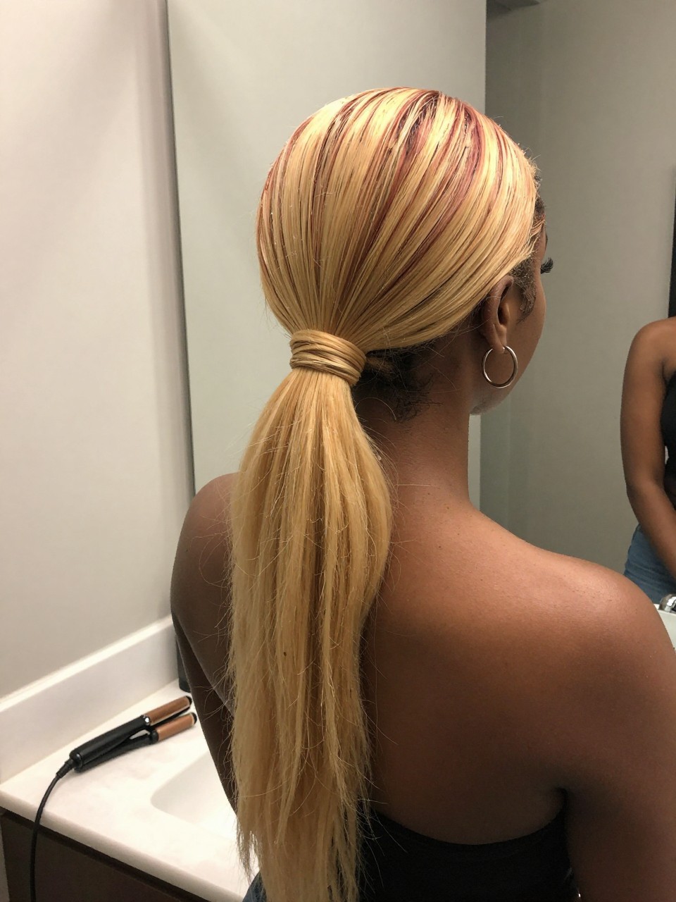 Photo of a black woman wearing a sleek mid ponytail with strawberry blonde lengths and bright blonde highlights, back-of-head view to highlight details, at a bathroom vanity mirror, soft indoor lighting, containing a small lived-in detail such as a flat iron on the counter and small hoop earrings peeking through, latest iPhone photo quality.