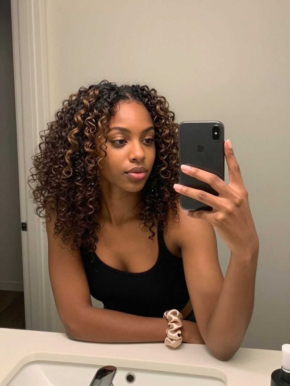 Photo of a Black woman wearing a defined natural coil out with mocha highlights throughout the curls, candid mirror selfie, at a bathroom vanity mirror, soft indoor lighting, containing a small lived-in detail such as a curl gel tube on the counter and a satin scrunchie on the wrist, latest iPhone photo quality.