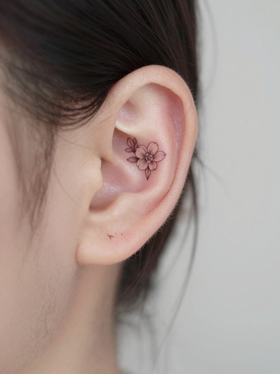Image Prompt: "Macro shot of a tiny jasmine flower tattoo behind the ear, fine line black ink floral design with small soft petals and light shading, realistic skin texture, a few loose strands of hair framing the placement, minimal feminine styling, tight crop, soft clean background, tattoo-centered inspiration image (no watermarks on images)"